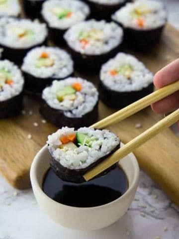 Ein Sushi wird mit Essstäbchen in Sojasauce gedopt mit mehr Sushi im Hintergrund