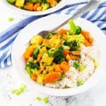 Gemüsecurry mit Reis in einem weißen Teller mit einer Gabel auf einer Marmorplatte mit einem blau weiß gestreiften Tischtuch im Hintergrund