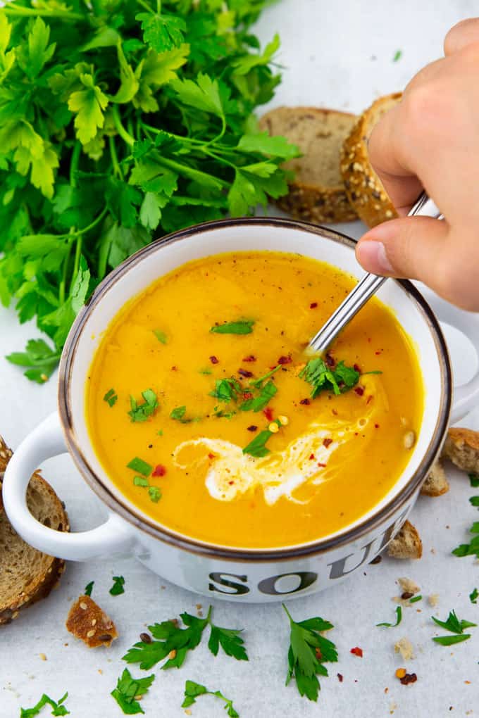 eine Suppenschüssel mit Kürbissuppe auf einer Marmorplatte mit einer Hand, die mit einem Löffel die Suppe löffelt