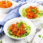 Zwei weiße Teller mit Zucchini Spaghetti und Tomatensauce auf einer Marmorplatte mit einer Gabel an der Seite