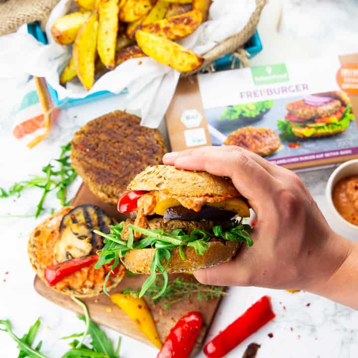 eine Hand, die einen veganen Burger hält mit Pommes und Grillgemüse im Hintergrund 