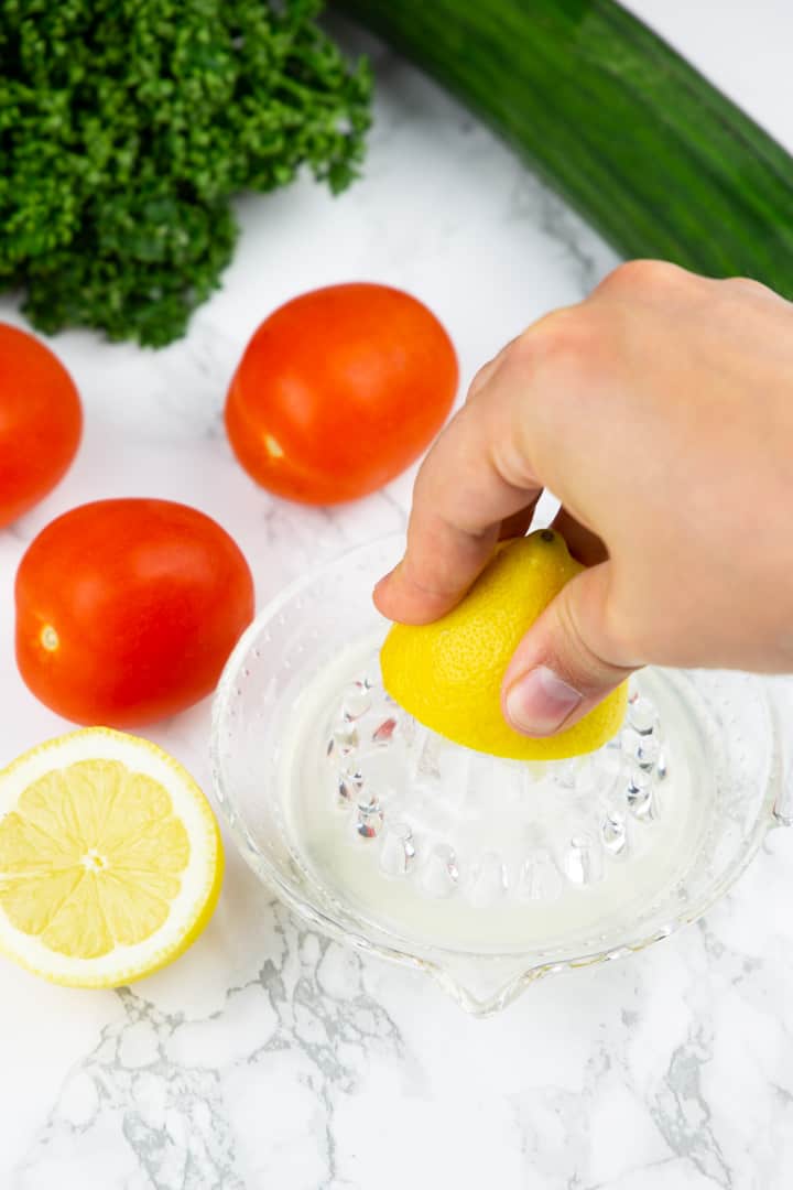 eine Hand, die eine Zitrone mithilfe einer Zitronenpresse aus Glas auspresst mit einer Gurke und Tomaten im Hintergrund 