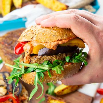 eine Hand, die einen veganen Burger hält mit Pommes und Grillgemüse im Hintergrund