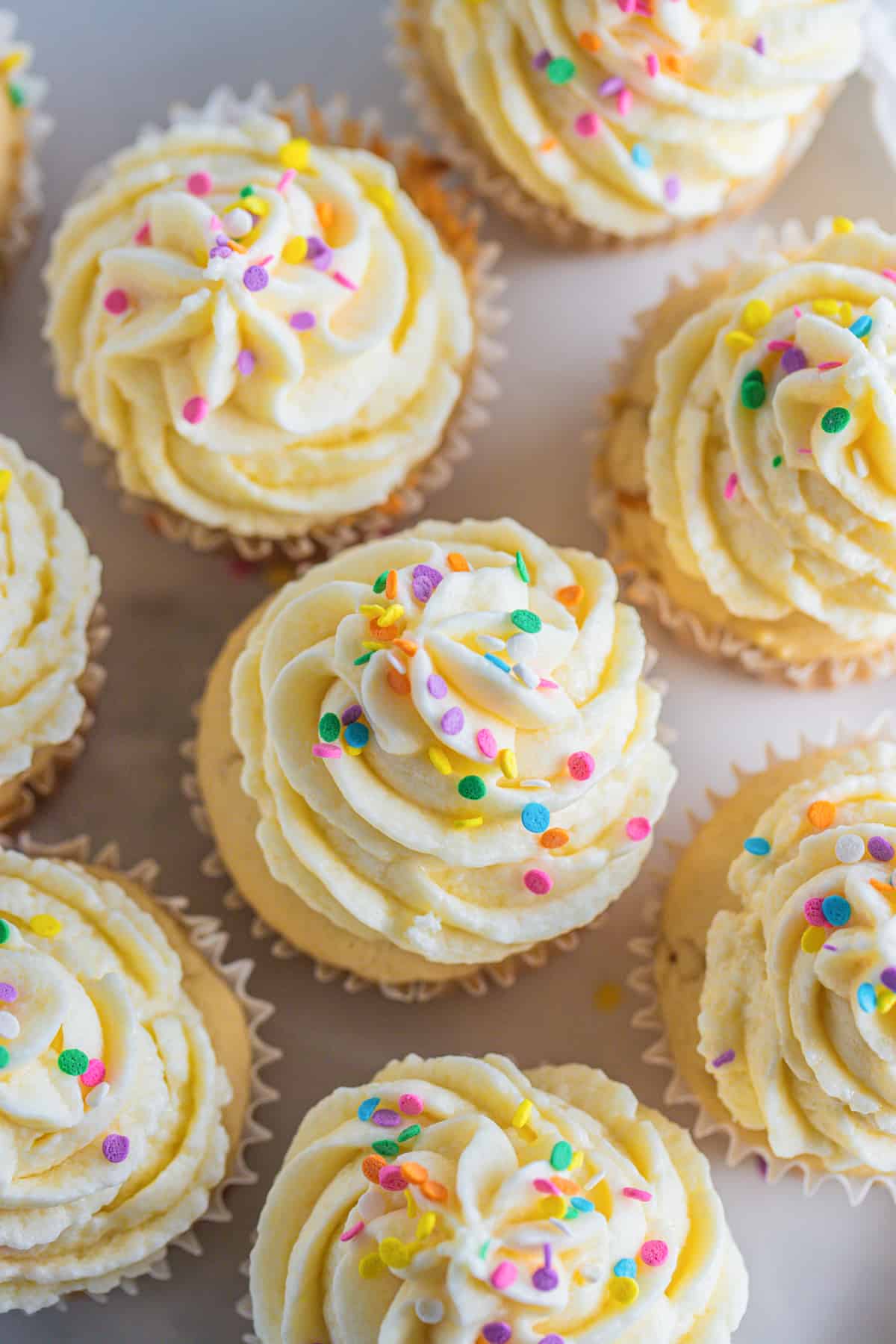acht vegane Cupcakes mit Vanille Frosting und Zuckerstreuseln auf einer hellen Arbeitsfläche 