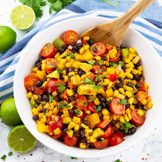 Mexikanischer Salat mit schwarzen Bohnen und Mais in einer weißen Schüssel mit Limetten und Korinder im Hintergrund