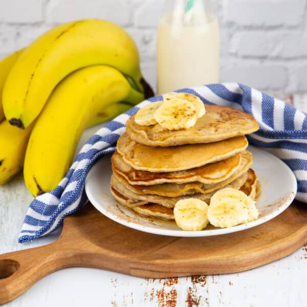 ein Stapel Bananen Pancakes auf einem weißen Teller mit Bananen im Hintergrund