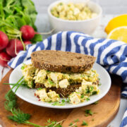 veganer Eiersalat auf dunklem Brot auf einem weißen Teller mit einem Bund Radieschen im Hintergrund