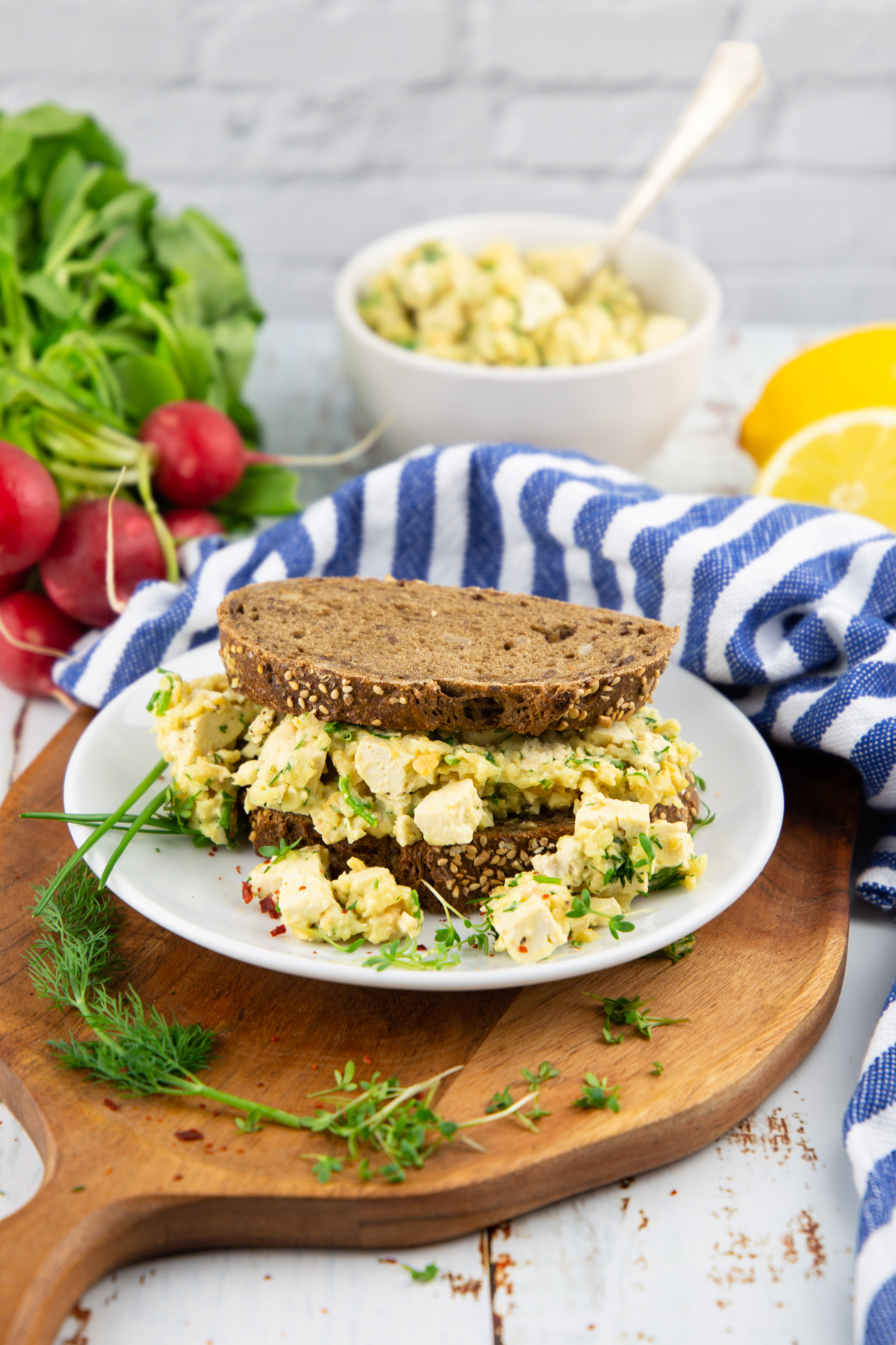 veganer Eiersalat auf dunklem Brot auf einem weißen Teller mit einem Bund Radieschen im Hintergrund 