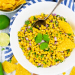 Mais Salsa in einer weißen Schüssel auf einem weißen Holzbrett mit Koriander und Limetten im Hintergrund