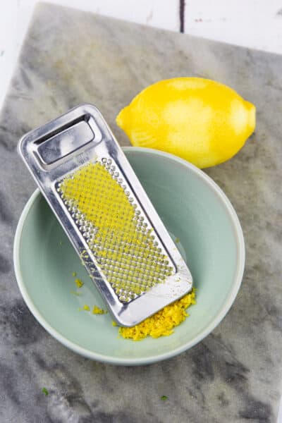 eine Zitrone mit einer kleinen Schüssel mit einer Reibe mit Zitronenabrieb auf einem Marmorbrett 