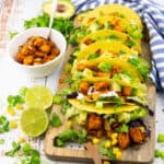 sieben Tacos gefüllt mit Tempeh, Salat und Avocado auf einem Holzbrett mit Limettenhälften und frischem Koriander daneben