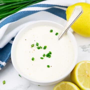 Vegane Sour Cream in einem weißen Schälchen auf einer Marmorplatte mit Zitronen und Schnittlauch daneben