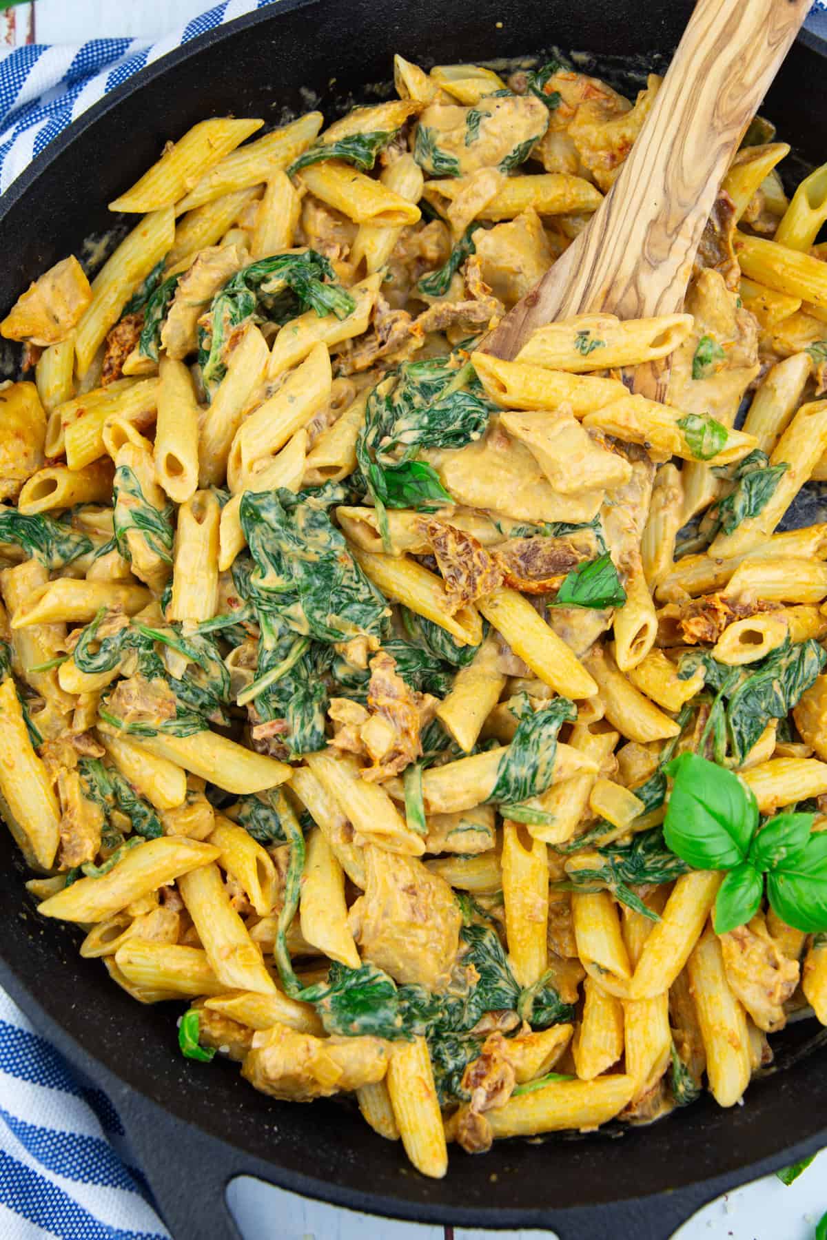 eine schwarze Pfanne mit Penne in einer cremigen Sauce mit Spinat auf einem blau-weiß gestreiften Tuch