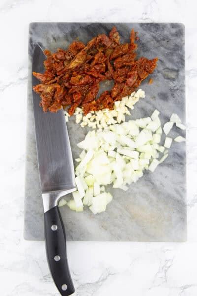 gehackte Zwiebeln, Knoblauch und getrocknete Tomaten auf einem Schneidebrett mit einem Messer daneben