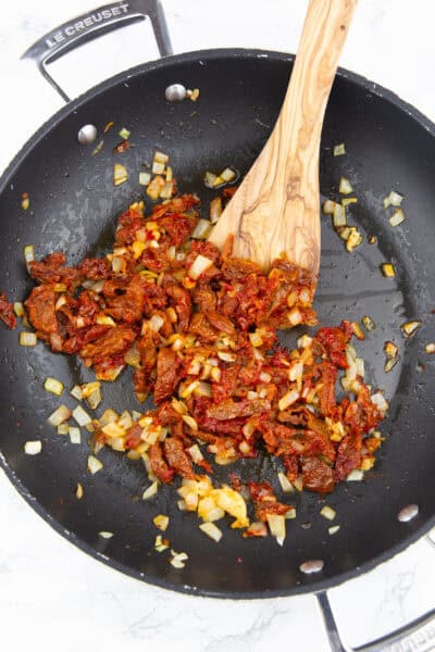 eine Pfanne mit getrockneten Tomaten, Zwiebeln und Knoblauch mit einem Holzlöffel