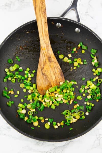 eine Pfanne mit angebratener Frühlingszwiebel und Knoblauch mit einem Holzspatel