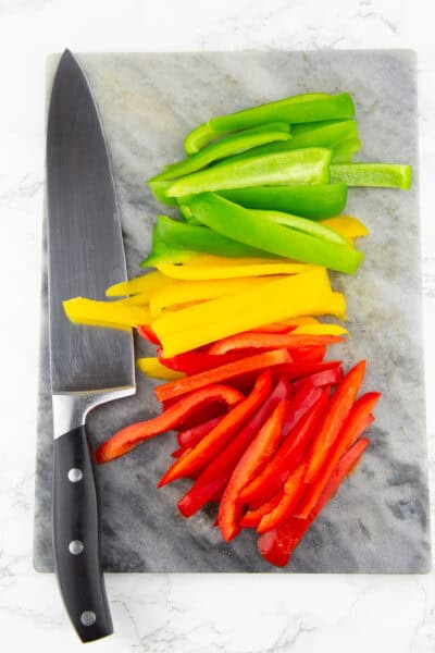 ein Marmorbrett mit in Streifen geschnittene bunte Paprika mit einem Messer daneben